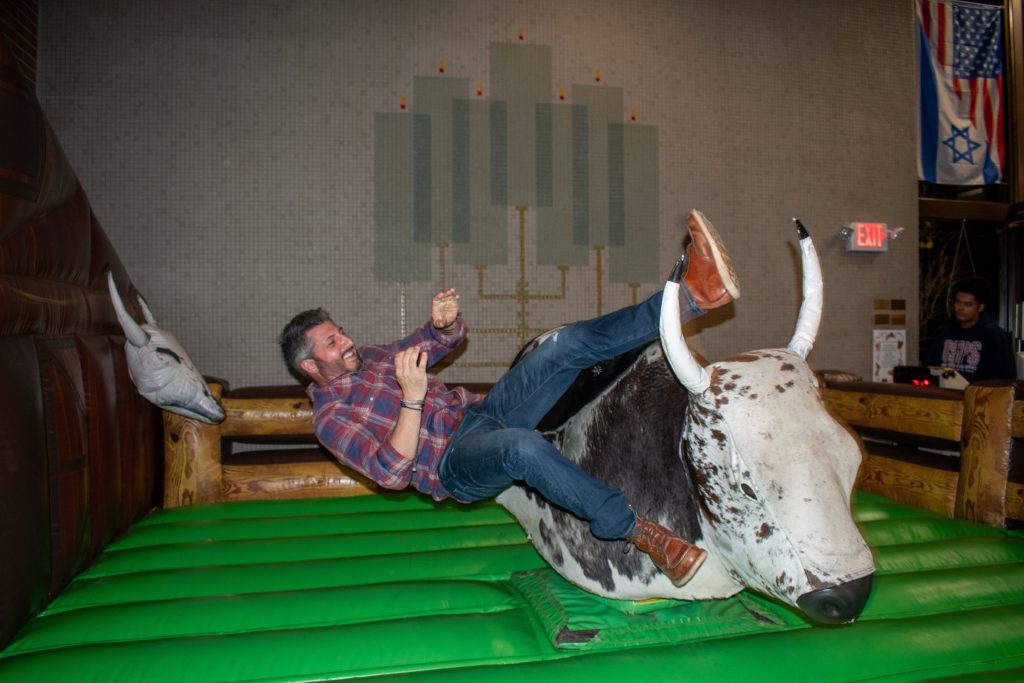 At the Challah Hoedown, a man in a plaid shirt and jeans is hilariously thrown off a mechanical bull indoors. His surprised expression and airborne legs amuse supporters nearby, as colorful flags adorn the lively background.