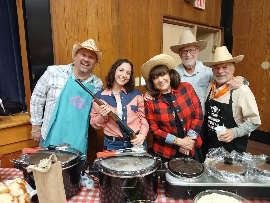 A group of five supporters stands behind a table filled with crockpots and dishes at the Challah Hoedown. They are smiling and wearing cowboy hats and casual western attire. One person holds a novelty rifle, adding to the festive indoor gathering or event.