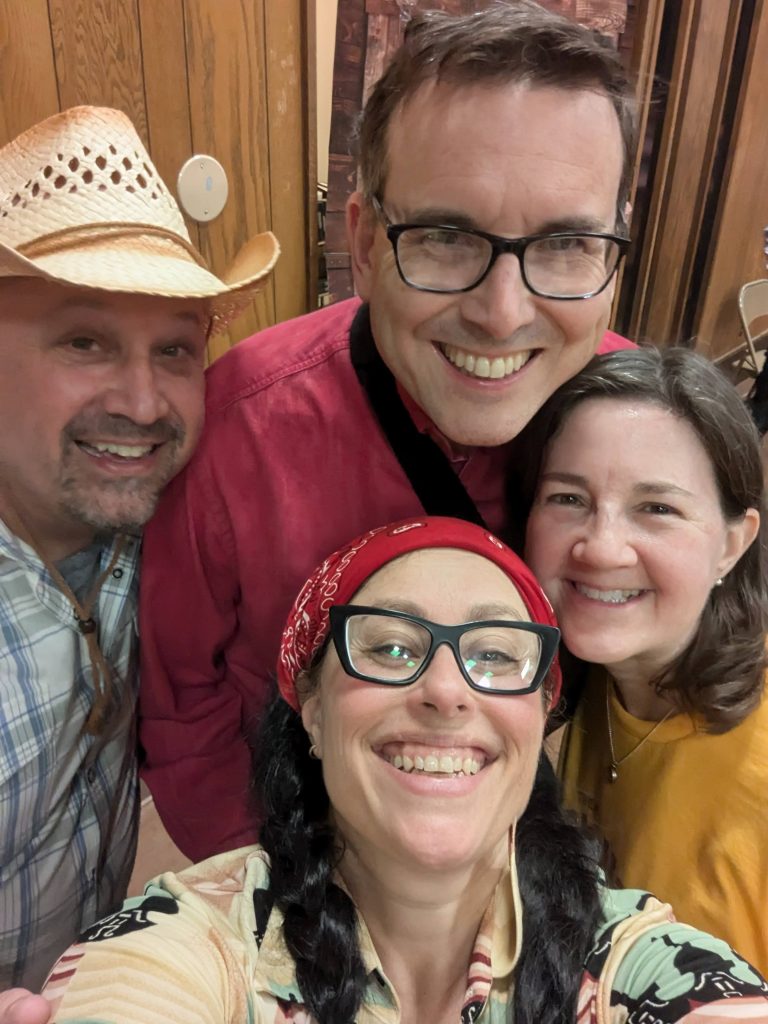 Four smiling friends snap a selfie indoors, celebrating with camaraderie and gratitude. Two sport classic glasses, while one dons a straw hat and another a red bandana. Perhaps this joyful snapshot is from the lively Challah Hoedown, where supporters gather in the warm wooden-paneled room.