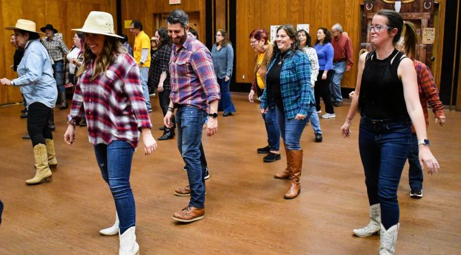 A group of Challah Hoedown supporters is line dancing in a wooden-floored room. They wear casual, Western-style outfits like plaid shirts, jeans, boots, and hats. With smiles all around, they create a lively and fun atmosphere.