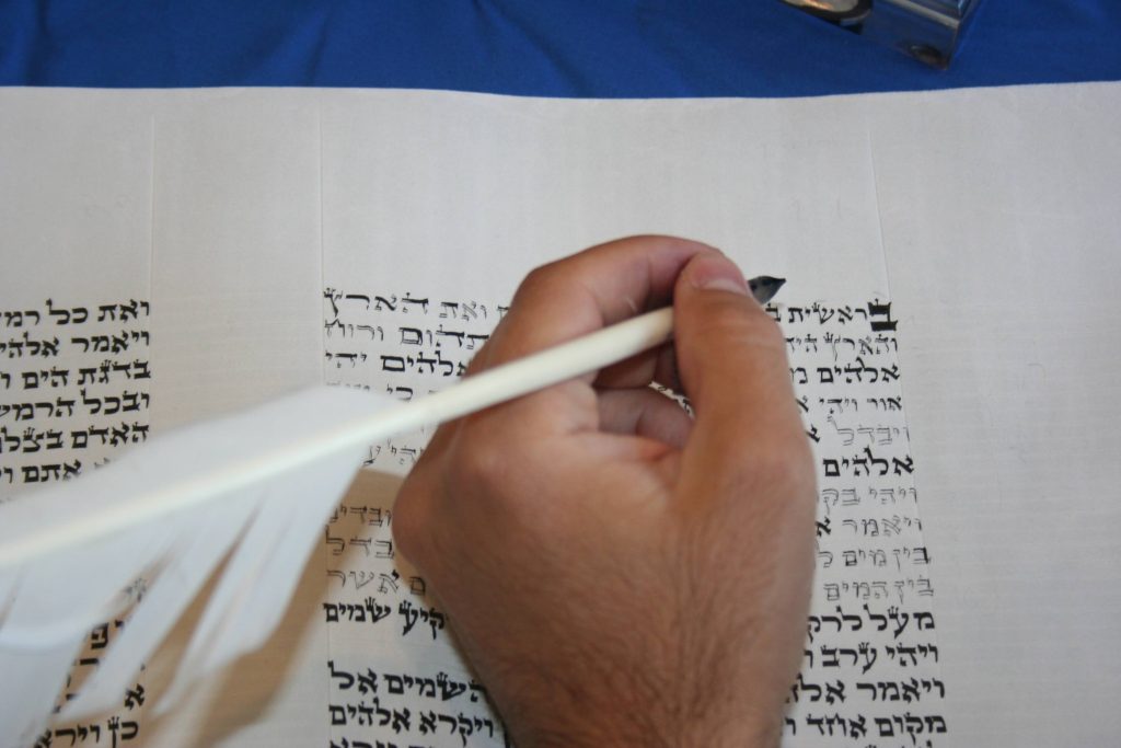 A person is meticulously writing Hebrew script on a scroll using a quill pen. The background is a blue cloth, and parts of the scroll reveal inked Hebrew text, possibly from the Torah. The hand in focus highlights the traditional writing process during this religious year.