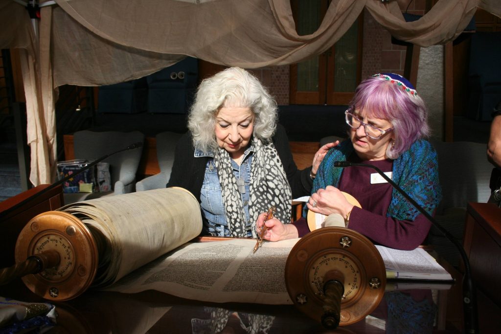 Two women sit together, closely examining and pointing at an open scroll with Hebrew text. Under the softly lit canopy, one wears a black and white scarf while the other, with purple hair and glasses, ponders over the Torah. It's a serene moment in this special Year of the Torah.