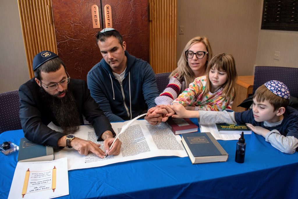 A bearded man with glasses guides a family of four in writing on a scroll during the Year of the Torah. The family, including a woman, a man, a young girl, and a boy wearing a kippah, holds the pen together. Books and papers are scattered on the blue-covered table.