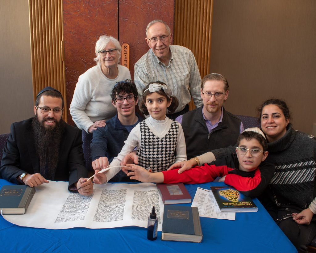 A diverse group gathers around a table with a large scroll and religious books. A child in a black and white outfit holds a quill, assisted by a man in a dark suit. They are engaged in a religious ceremony marking the Year of the Torah according to the Jewish calendar.